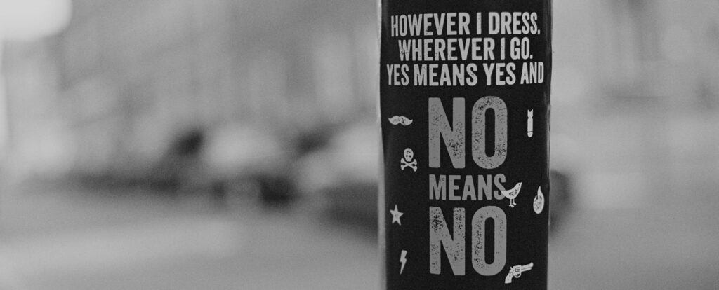 Schwarz-Weiß-Foto eines Straßenaufklebers an einem Laternenmast mit dem Text ‚However I dress, wherever I go, yes means yes and no means no', umgeben von kleinen Symbolen wie Totenkopf, Stern und Blitz.